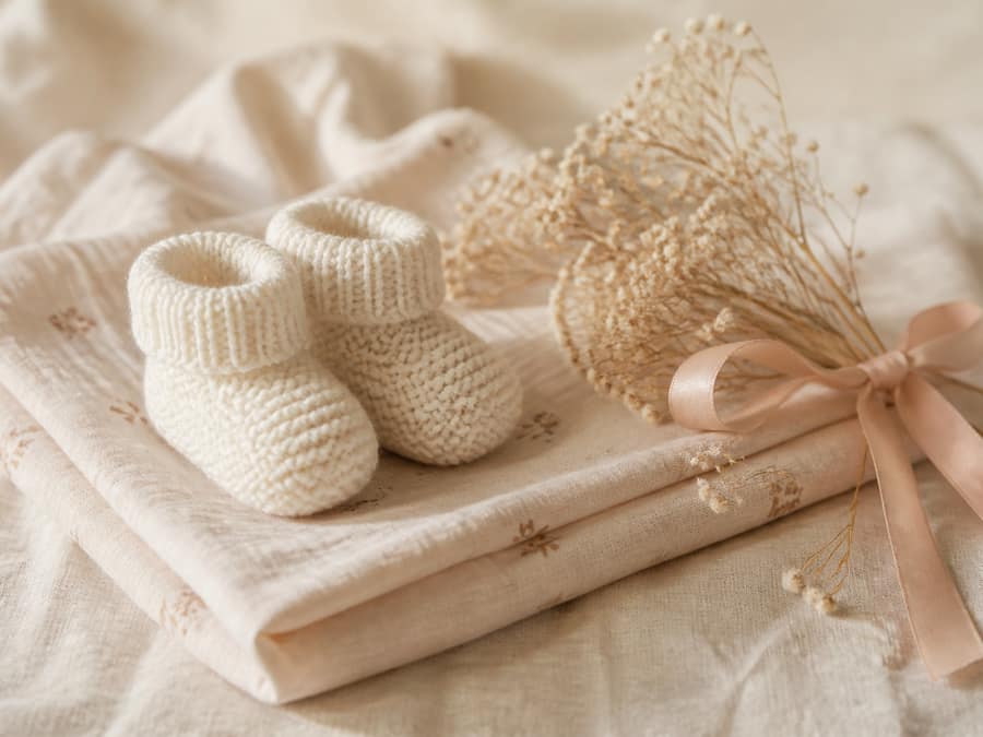 Knitted ivory baby booties on a folded muslin blanket with dried flowers