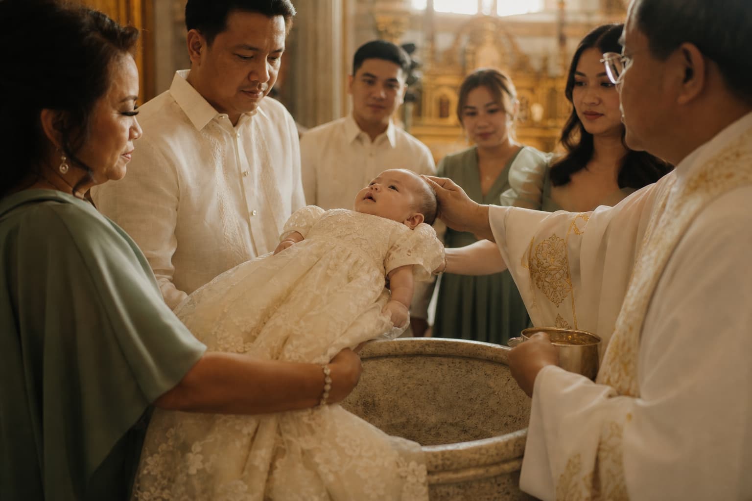 A baby in an ivory lace gown held above a stone baptismal font, godparents in cream barong and sage dresses gathered behind