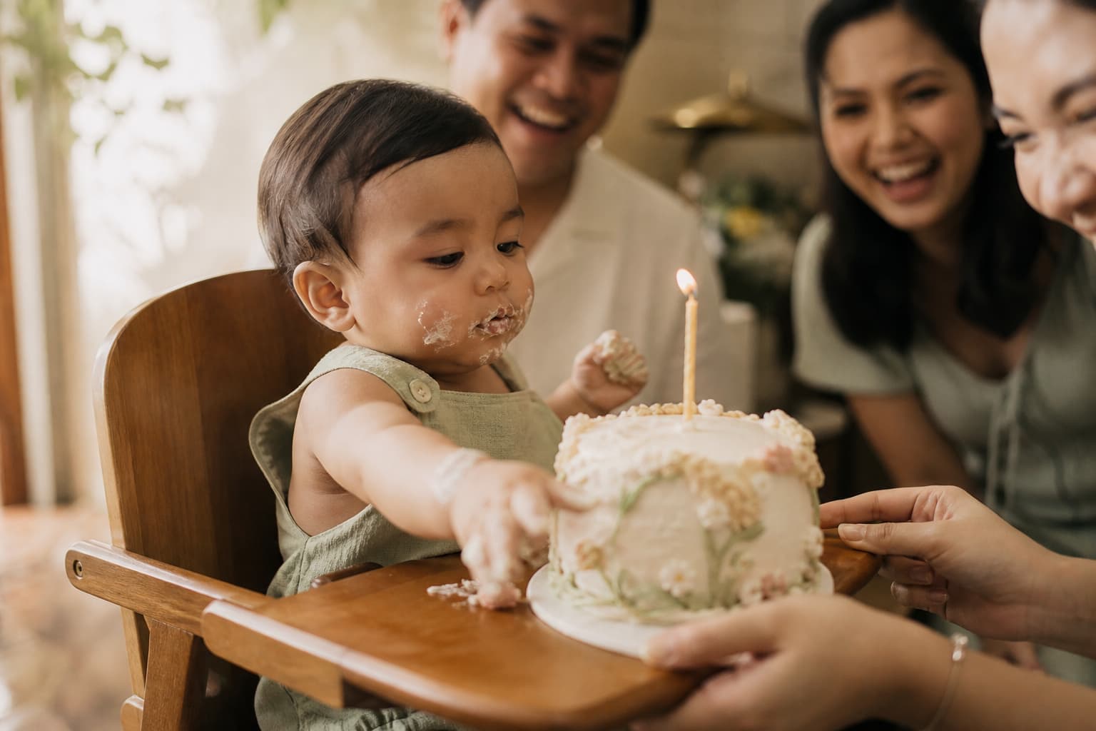 A one-year-old in a wooden high chair reaching for a small pastel smash cake with a single candle, family laughing behind