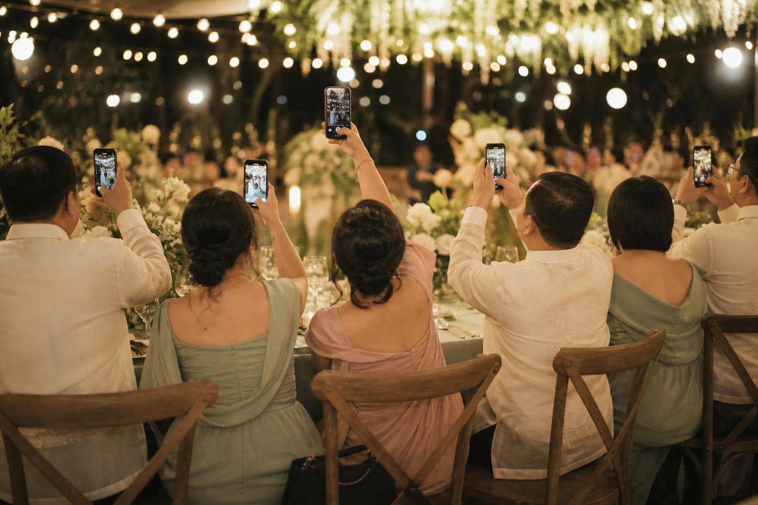 Wedding guests seen from behind at a long reception table, several holding phones up to photograph something ahead of them under string lights