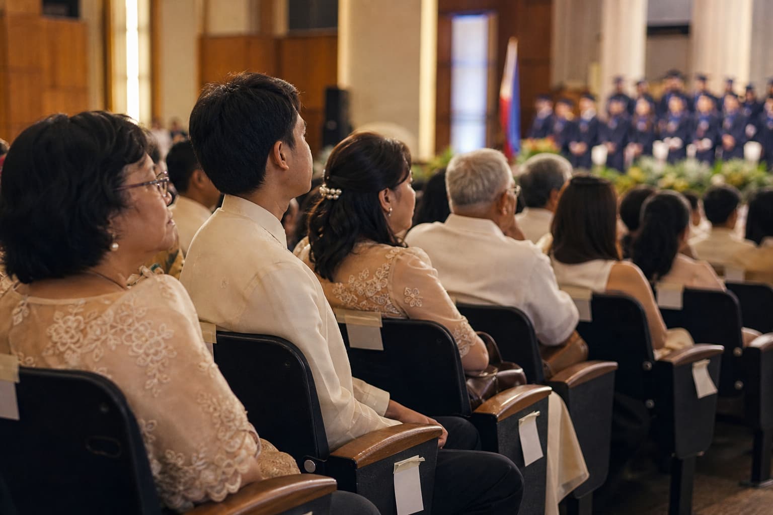 Rows of seated families in a Philippine university auditorium with small reserved cards on the seat backs, graduates in navy gowns beyond