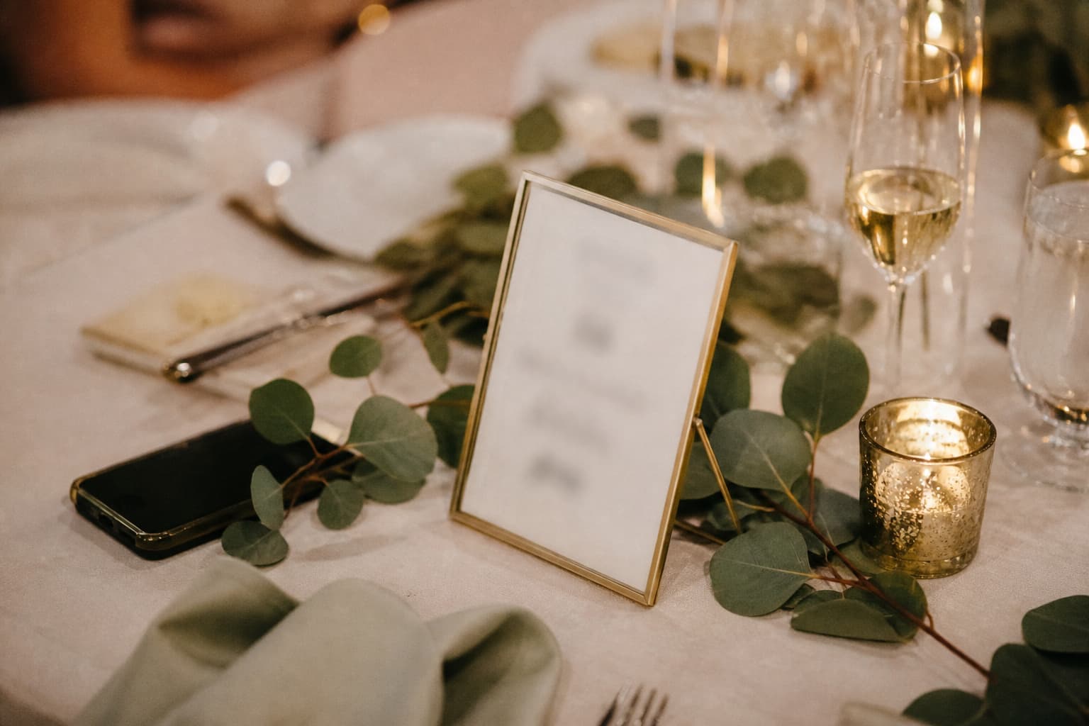 A small brass-framed table sign on a reception table surrounded by wine glasses, a votive candle and eucalyptus