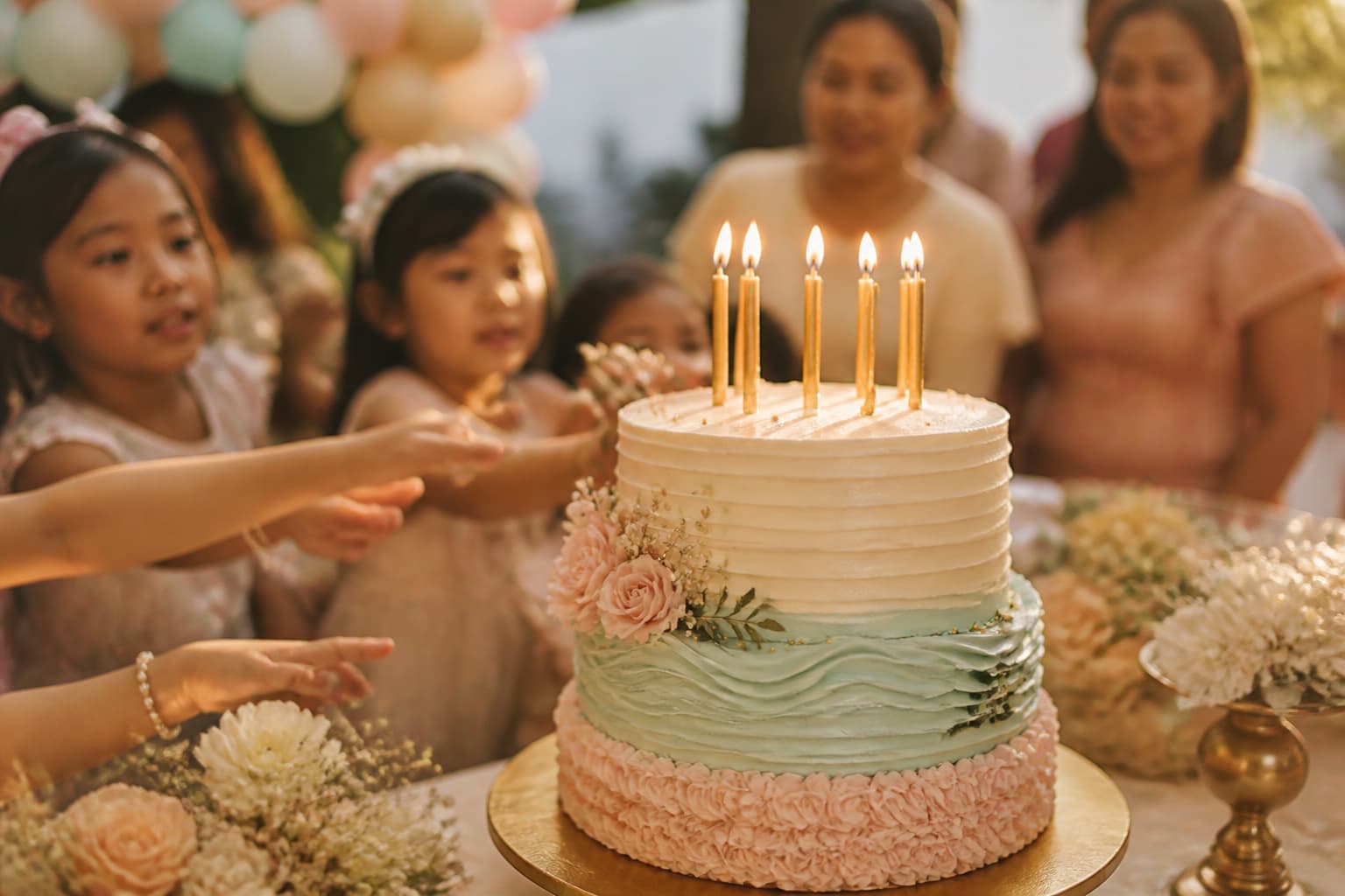 A pastel layer cake with seven lit candles on a decorated party table, children's hands reaching in from the edge of frame